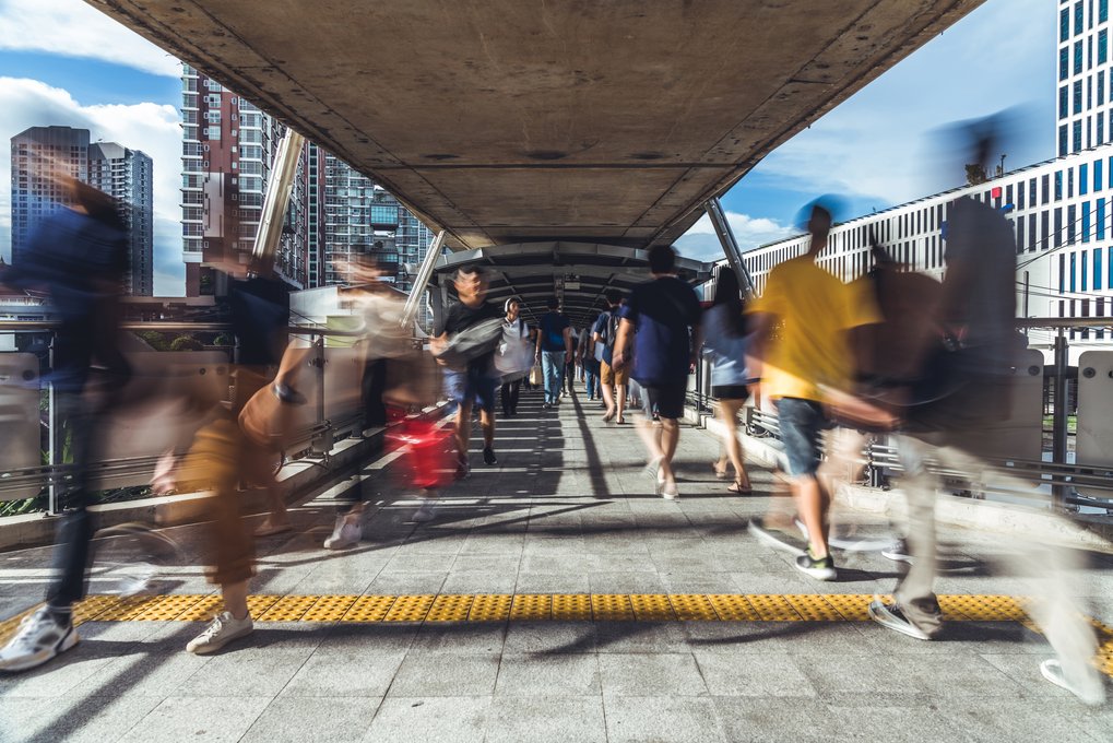 Pedestrians on a covered walkway, surrounded by high-rise buildings and blue skies in the background.